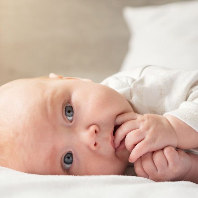Baby girl lies on side on big white parent bed. Newborn child with grey eyes and plump cheeks lies in bedroom putting his fingers in his mouth and sucking his fingers, sunlight